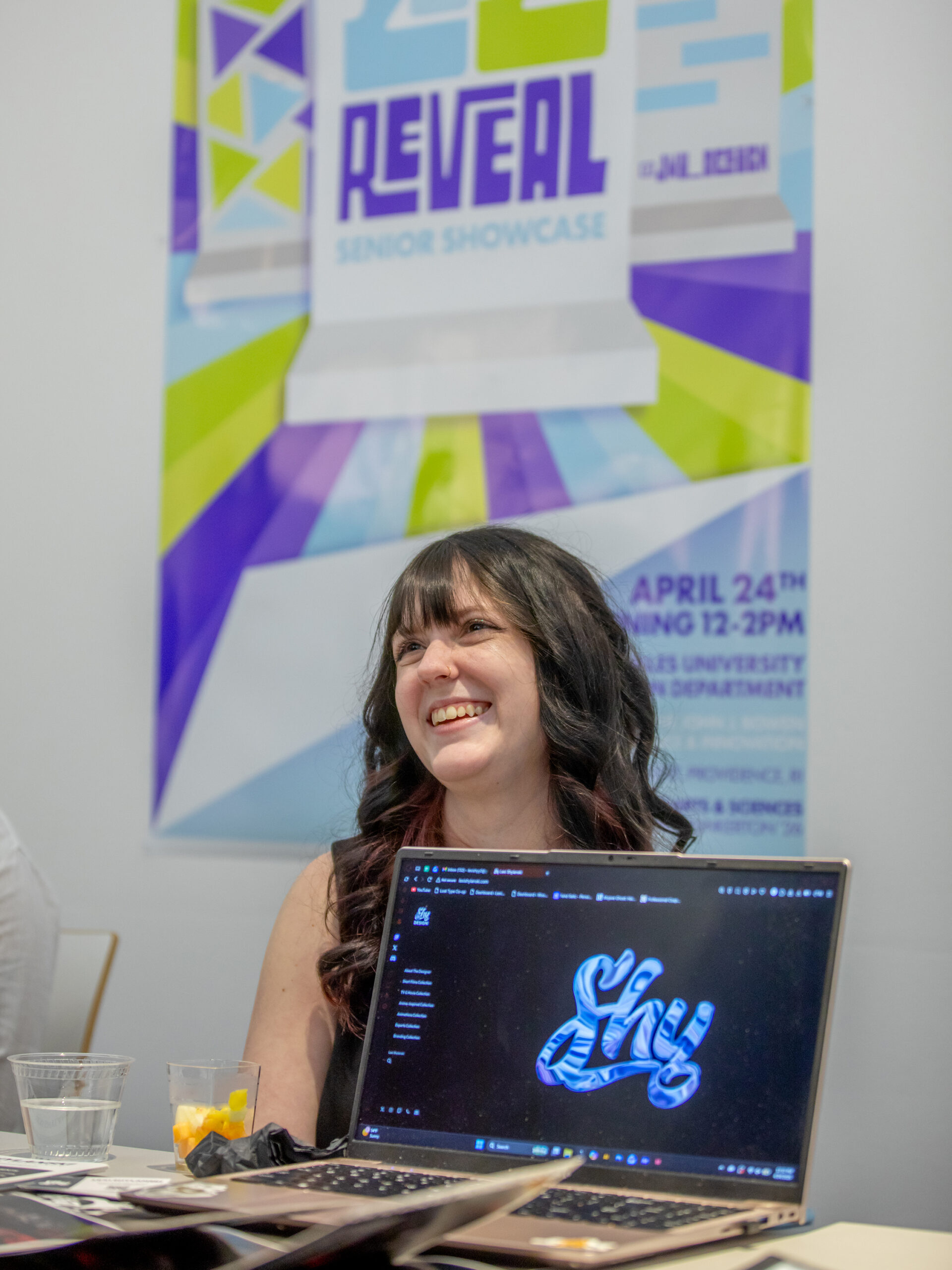 A person smiles while seated at a table with a laptop displaying a stylized "Sky" logo, in front of a poster for the "Class of 2026 Reveal Senior Showcase."