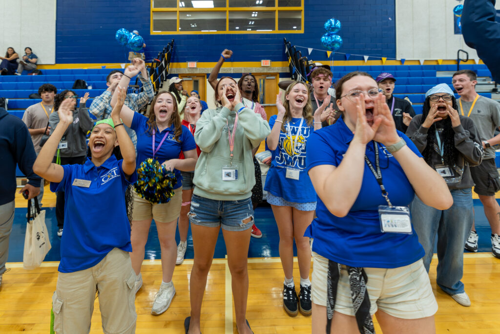 A group of young people in a gymnasium cheer enthusiastically, some with arms raised and others clapping, while wearing blue shirts and lanyards.