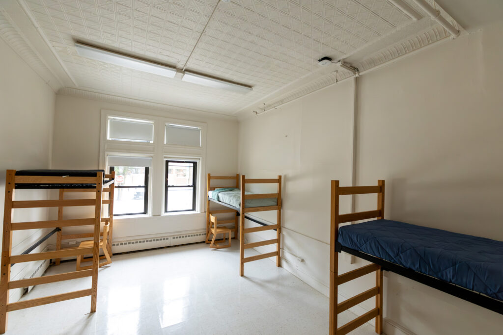 A clean, empty dormitory room with three wooden bunk beds, one with a blue mattress, and a window letting in natural light.