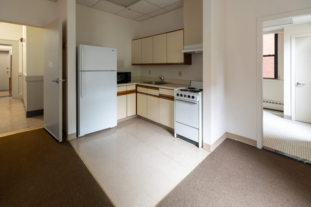 A clean, minimalist kitchen with white appliances and beige cabinets, viewed from a hallway with carpeted floors and open doorways.
