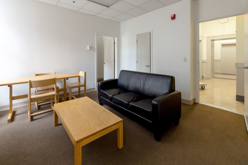 A minimalist waiting room features a black leather sofa, wooden tables and chairs, and an open doorway leading to a hallway.