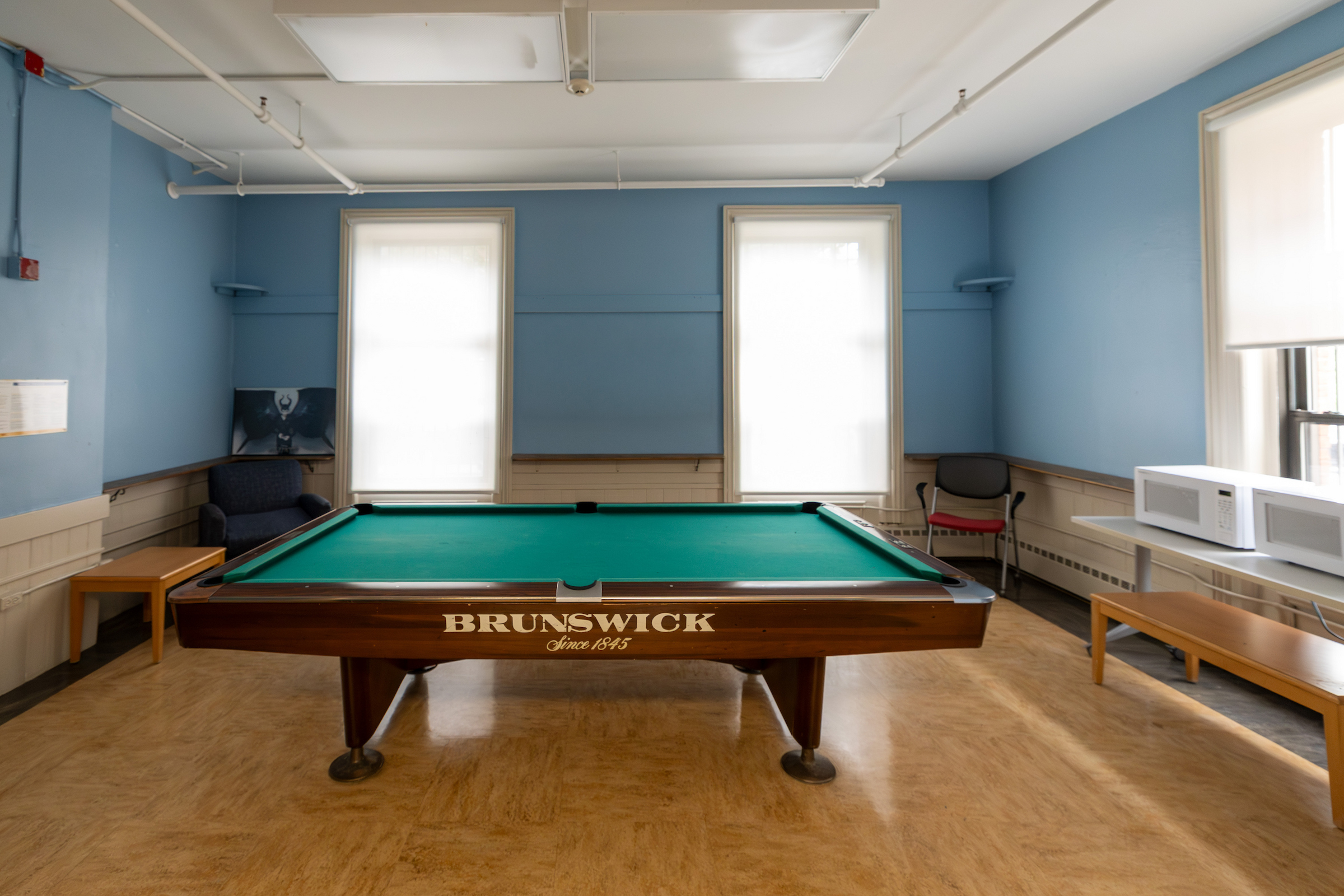 A Brunswick pool table sits in a room with blue walls, wooden floors, and large windows, surrounded by seating and a microwave.
