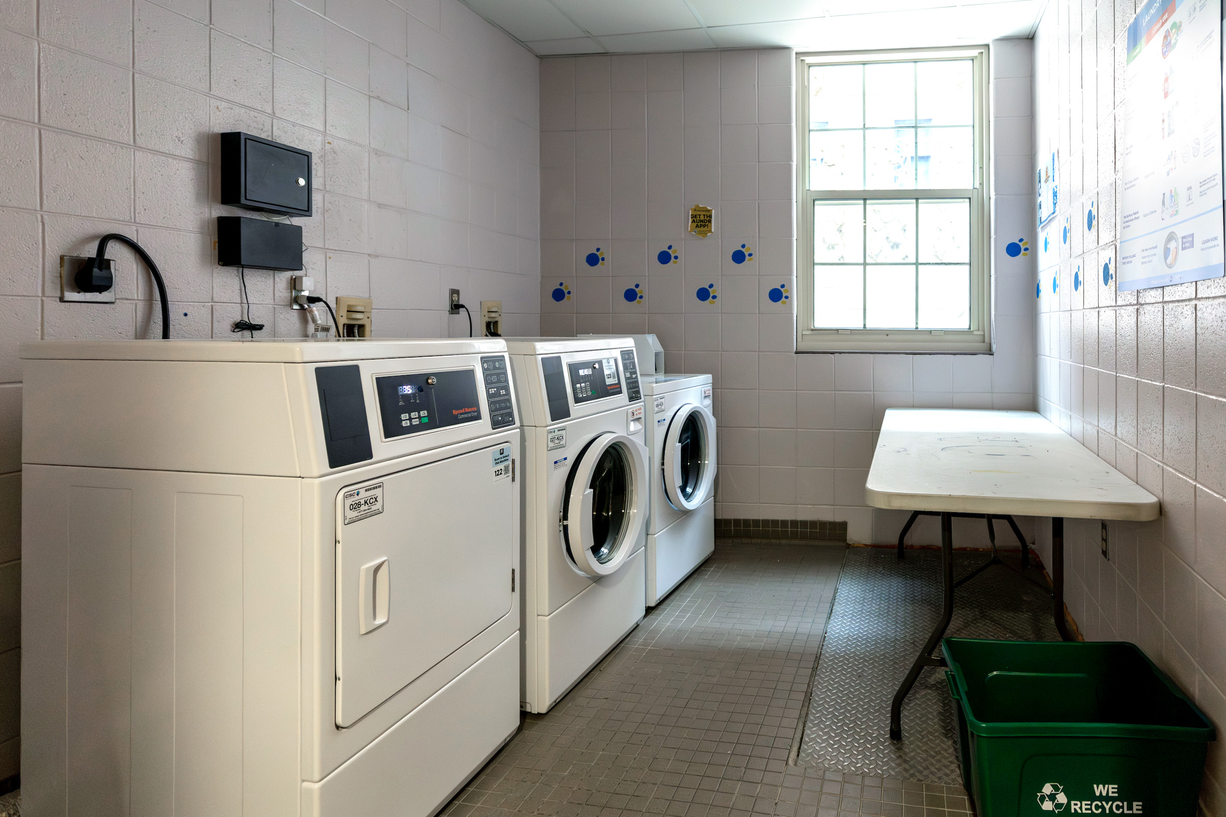 A row of white laundry machines lines a tiled room with a window and a green recycling bin.