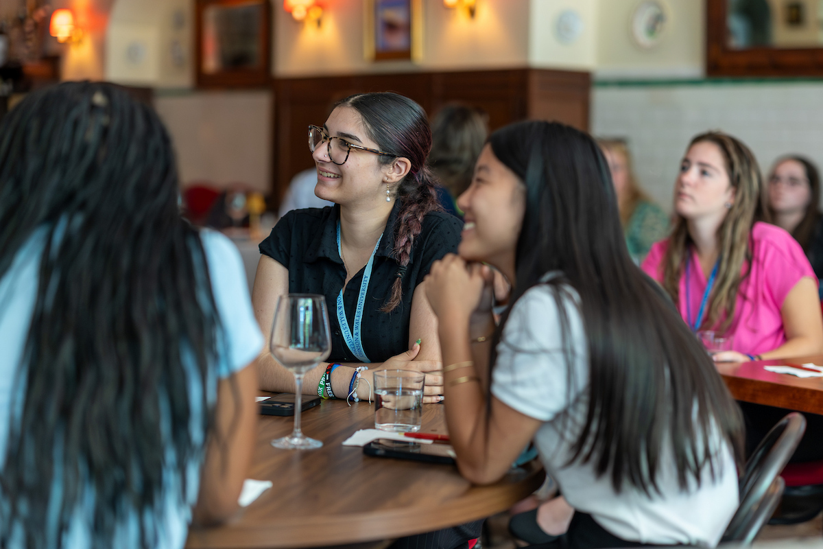 A group of young people are seated at a table in a warmly lit restaurant, engaged in conversation and smiling.