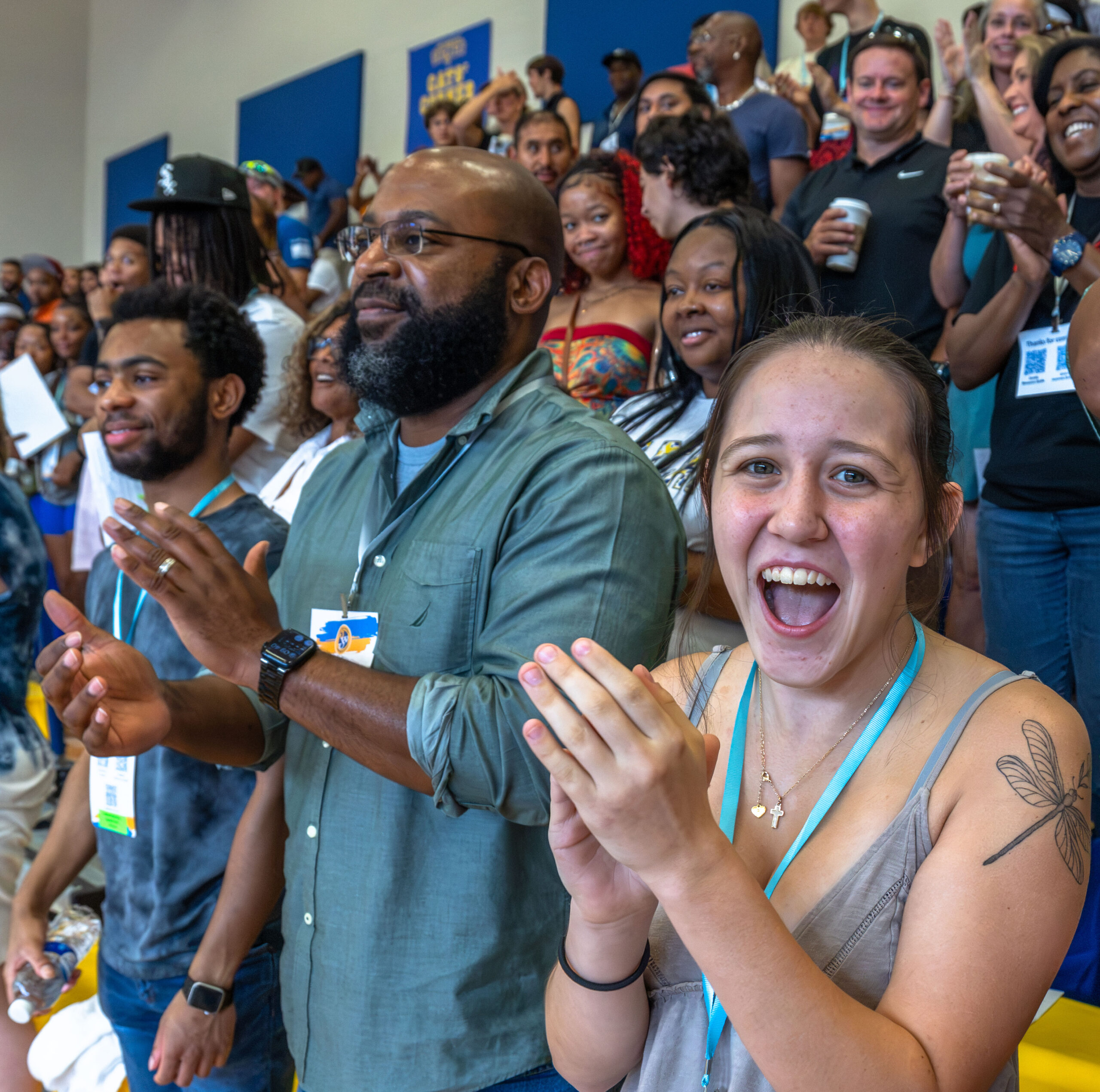 A diverse group of people in a gymnasium applaud enthusiastically, with some holding awards or certificates.