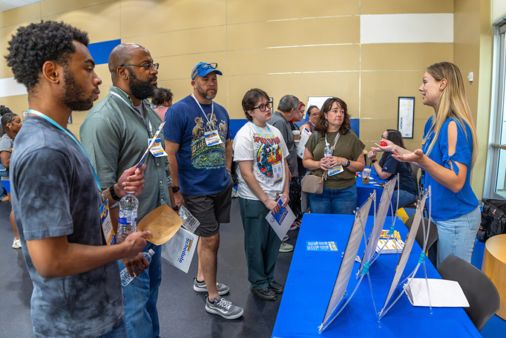 A group of individuals engages with a presenter at an informational booth during what appears to be a professional or educational event.