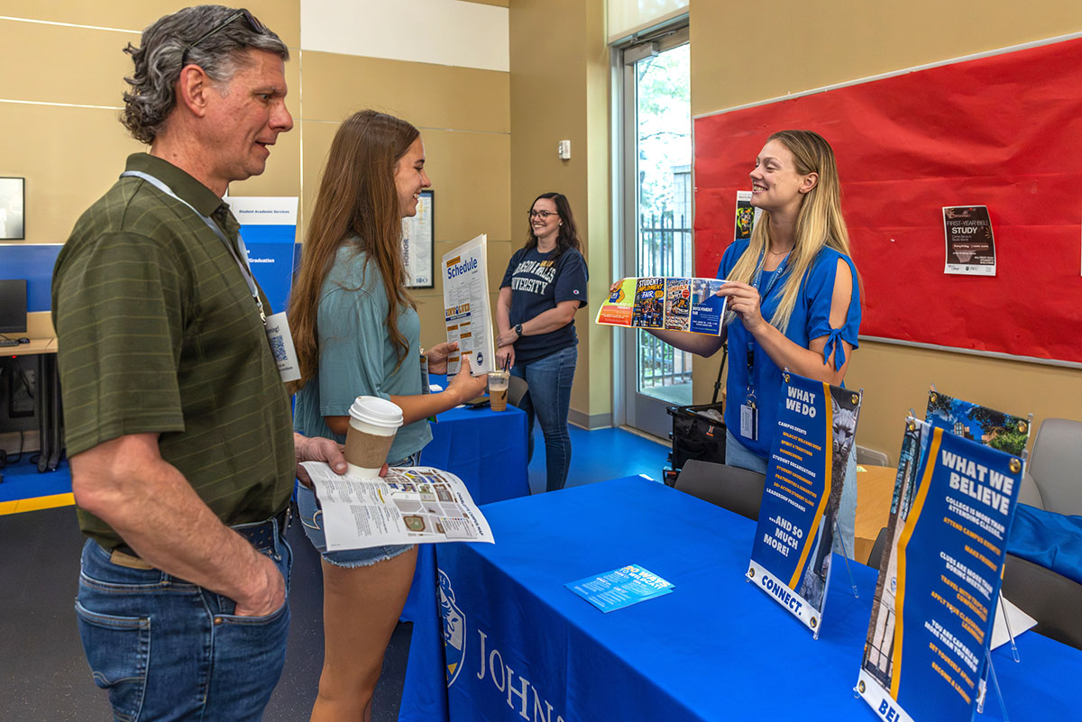 A new student and a parent stand at a blue table with promotional materials, engaging with a member of the cat team holding a brochure.