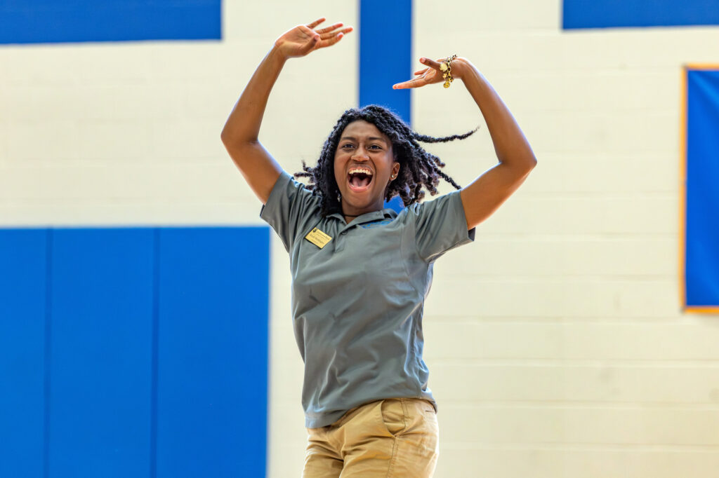 A person with braided hair, wearing a gray shirt and khaki pants, joyfully raises their arms with an open-mouthed expression in a gymnasium with blue and white walls.