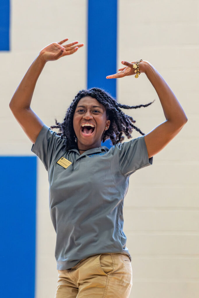 A member of the cat team, wearing a gray shirt and khaki pants, joyfully raises their arms with a big smile in a gymnasium.