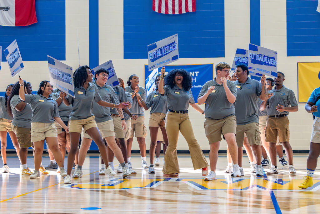 A group of young people in matching gray shirts and khaki shorts dance energetically in a gymnasium, holding signs that read "Royal Legacy" and "CIT Trailblazers."