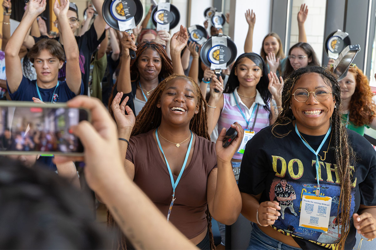 Orientation students joyfully raise their arms while one person takes a photo of the group in a brightly lit indoor space.