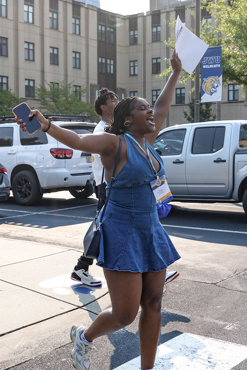A student joyfully holds up a paper while walking across a street on campus, with parked vehicles in the background.