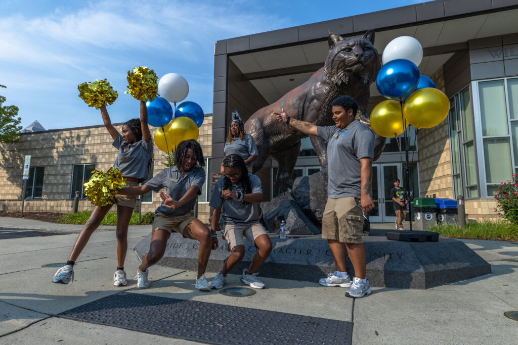 Cheerleaders and staff pose energetically around a large statue of a cougar, surrounded by colorful balloons, in front of a modern building.