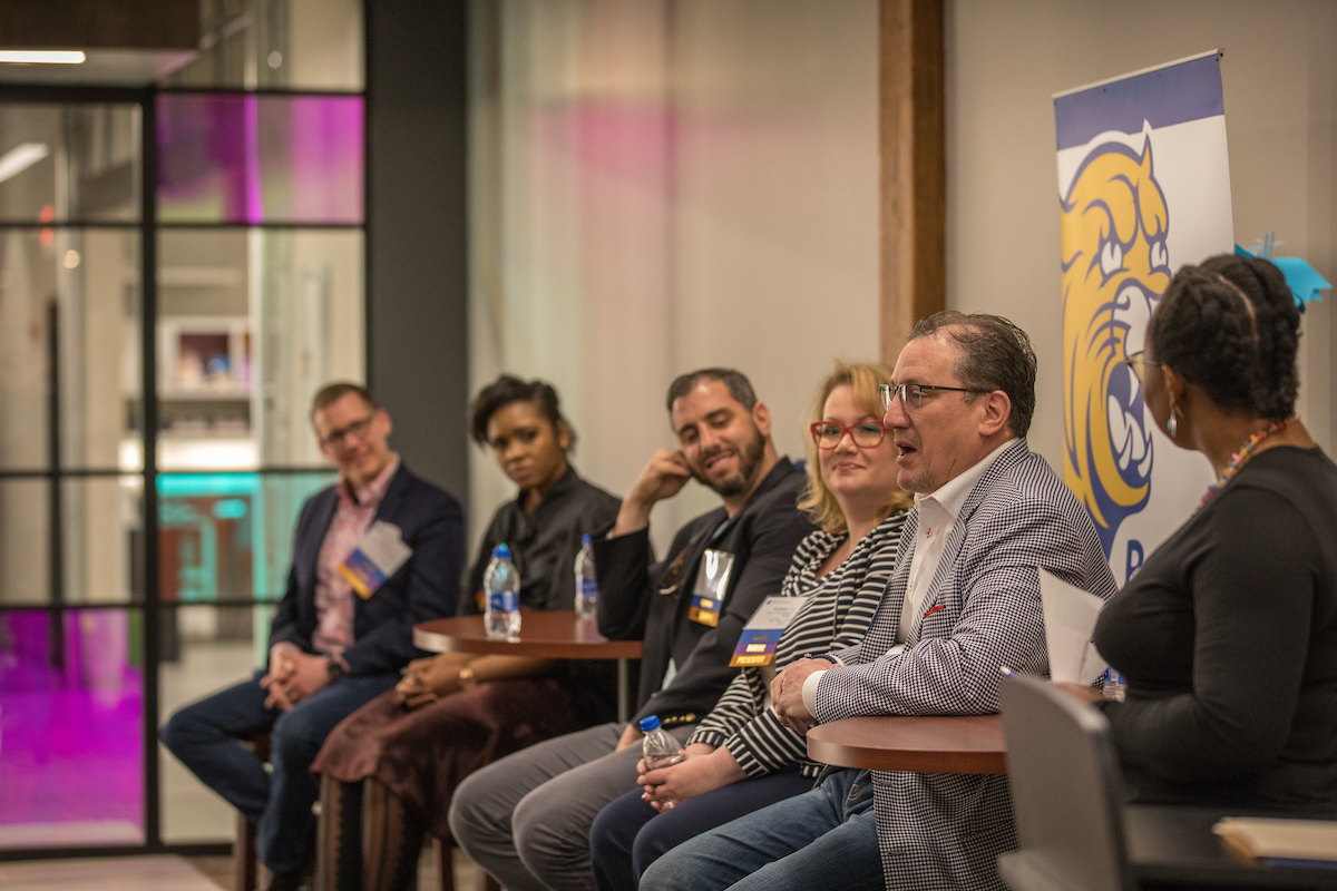A group of individuals sits in a row at a table during an event, with one person speaking while others listen, and a banner featuring a tiger mascot is visible in the background.