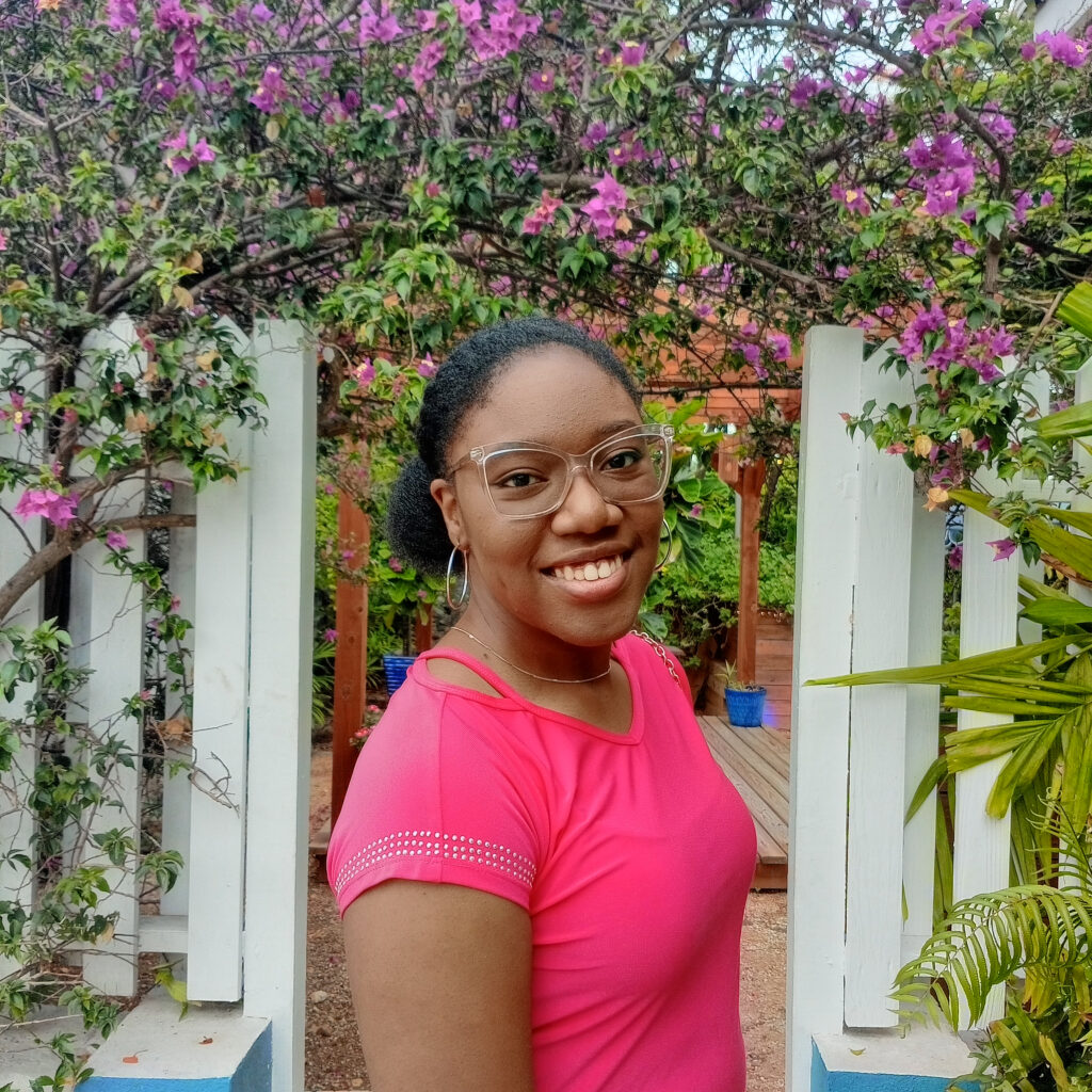 Rhema Blackman poses in front of a white fence with a tree full of purple flowers overhead.