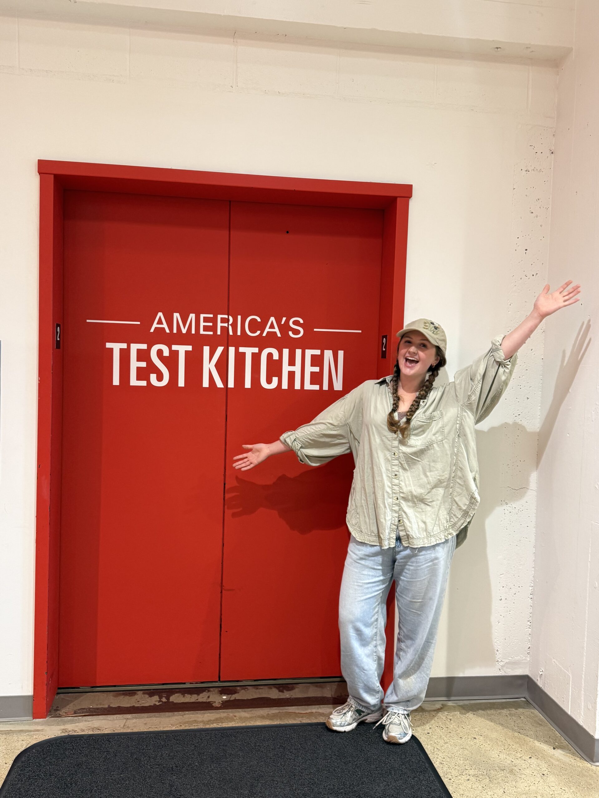 Avery Garmon stands with both arms outstretched beside the red America's Test Kitchen doors.