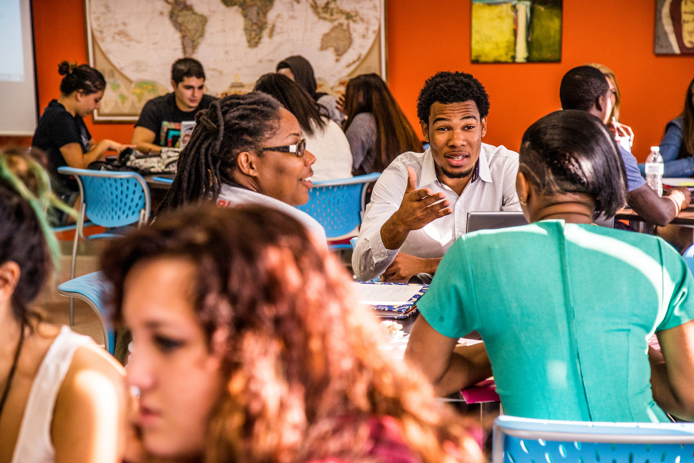 A diverse group of individuals engages in lively conversation around tables in a brightly lit classroom with a world map on the wall.