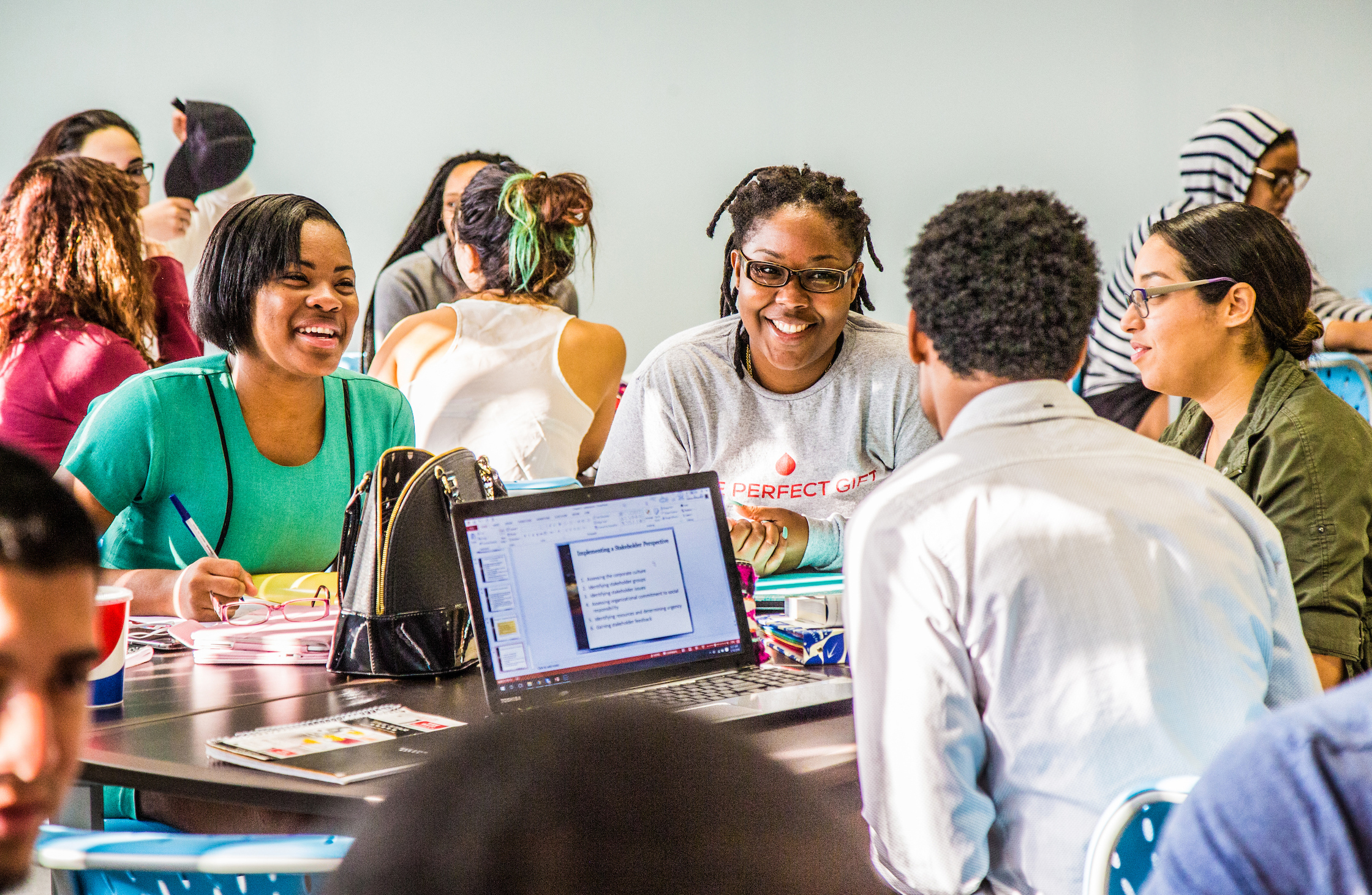 A group of people collaborate around a table in a classroom setting, with one person smiling while looking at a laptop displaying a presentation.