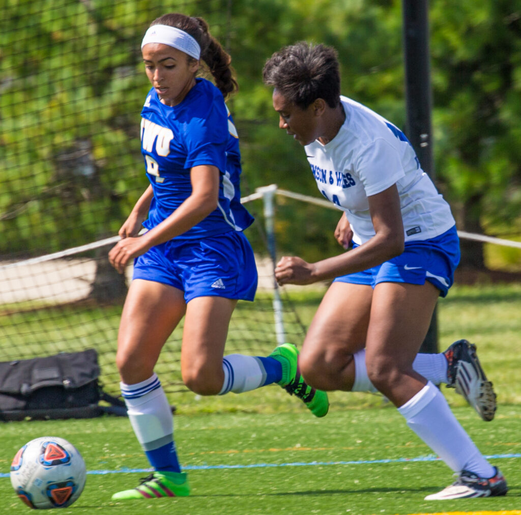 Two soccer players compete for the ball on a grass field, one in a blue uniform and the other in a white uniform.