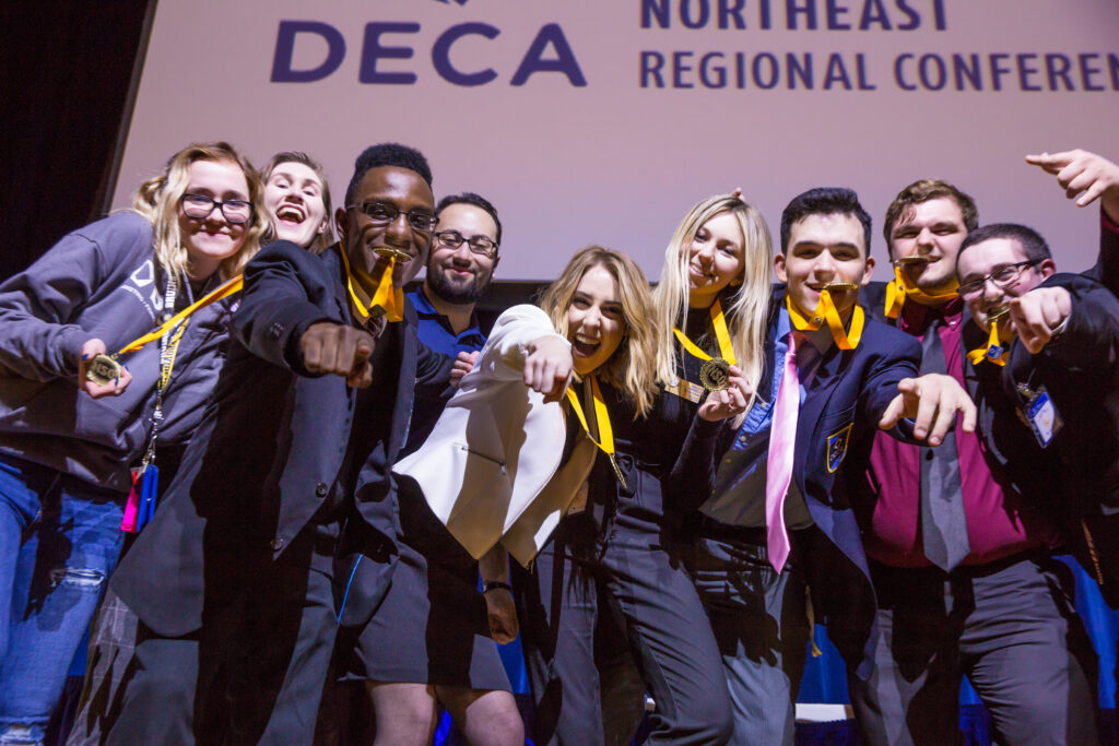 A group of young people celebrate at the DECA Northeast Regional Conference, proudly displaying their medals and pointing toward the camera.