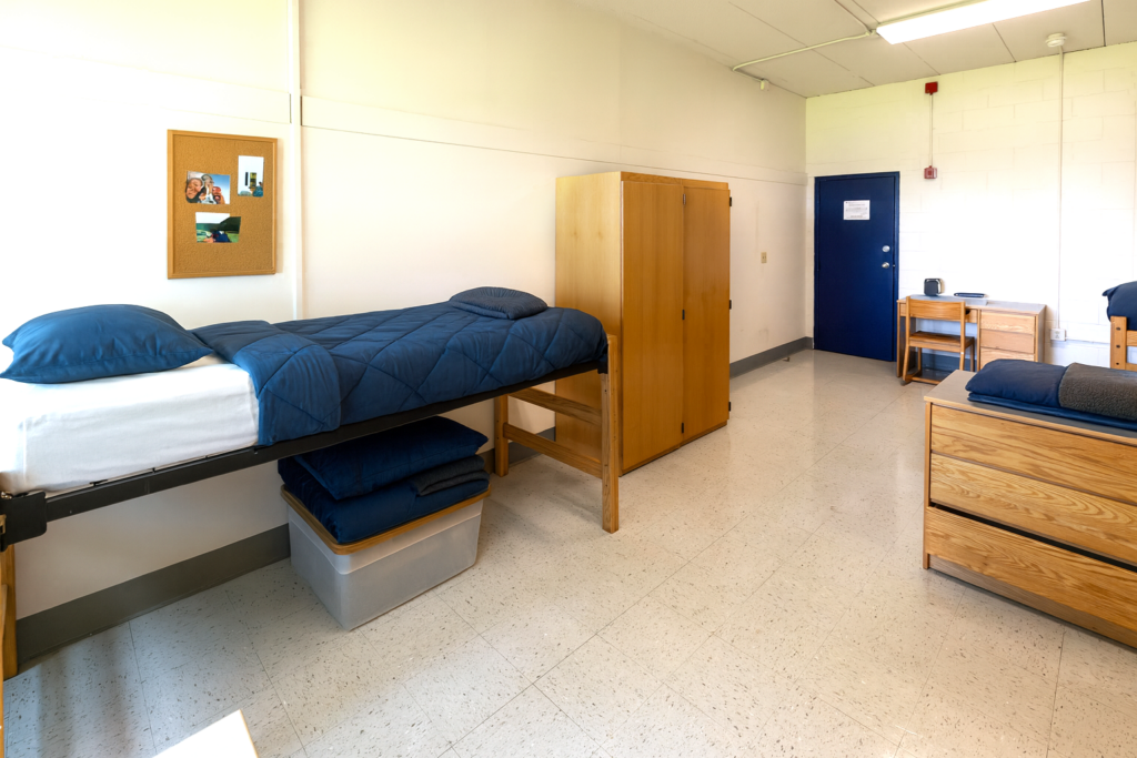 A dorm room with a twin bed, wooden desk, storage and a window between the bed and wall.