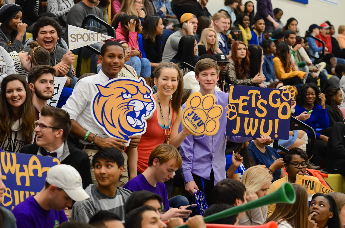 A lively crowd of students cheers at a school event, holding signs and a large mascot cutout for that says let's go J W U.
