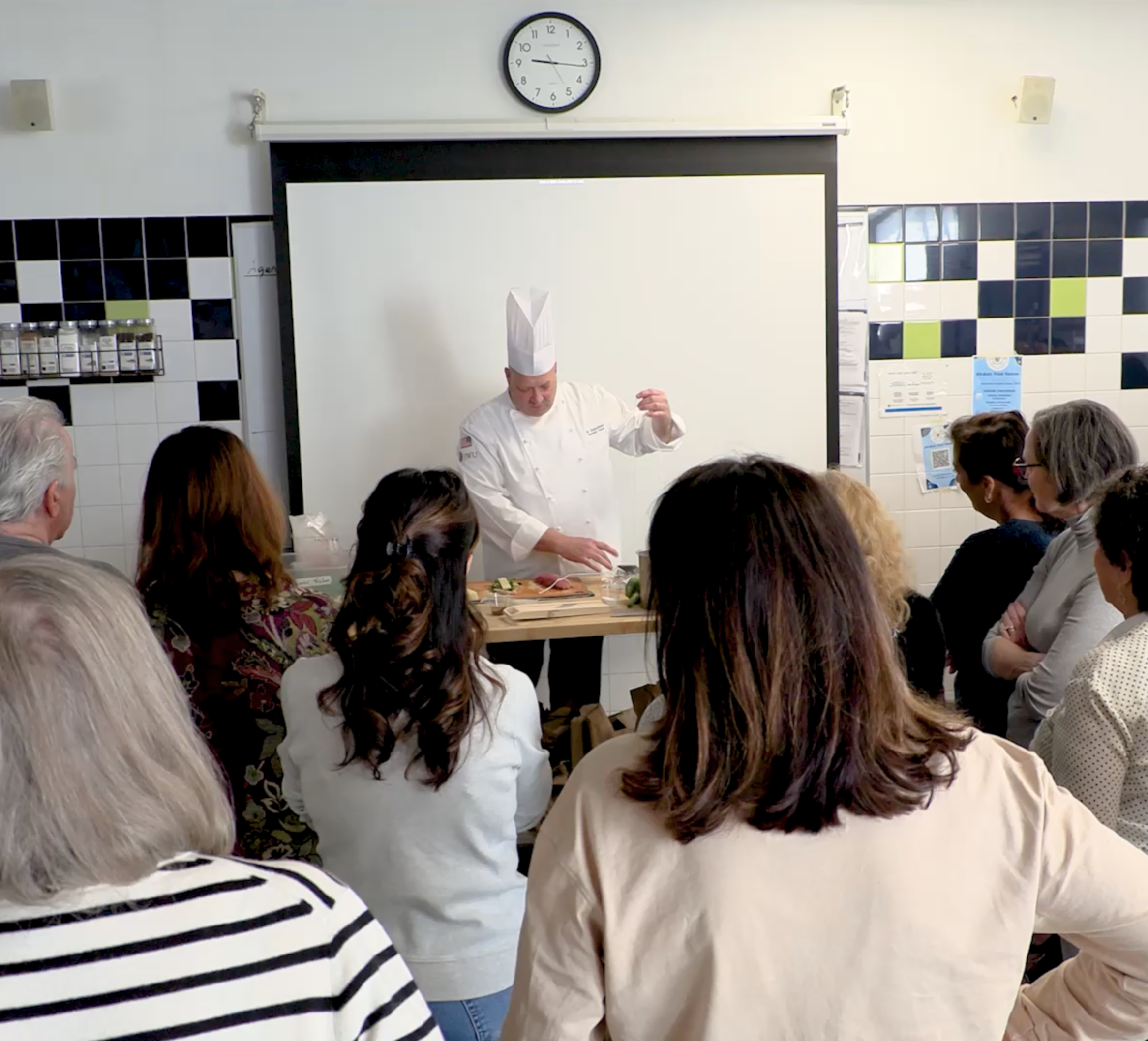 A group of adults stand around a demonstration table as a chef prepares ingredients