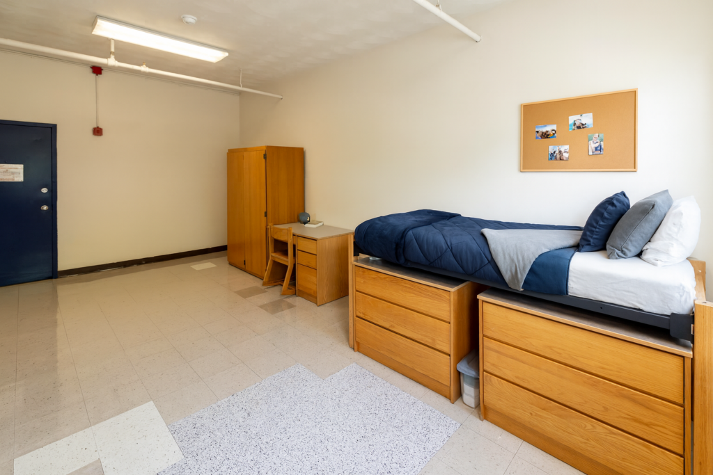 A dorm room with a twin bed wooden desk, storage and a chair.