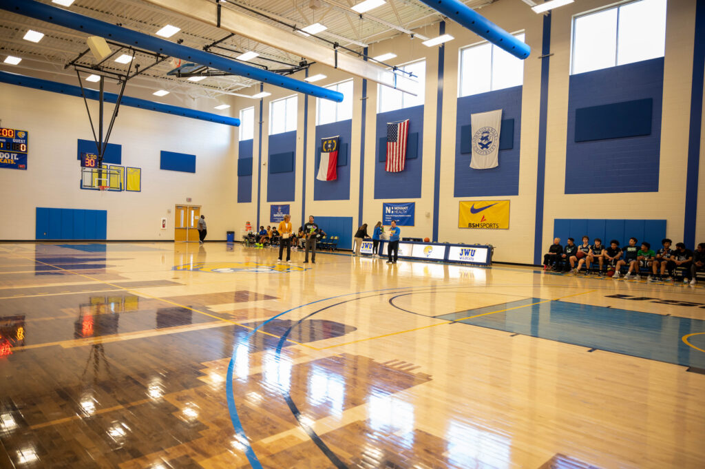 A polished wooden basketball court in a brightly lit gymnasium with players and staff gathered near the sidelines, surrounded by banners and flags.