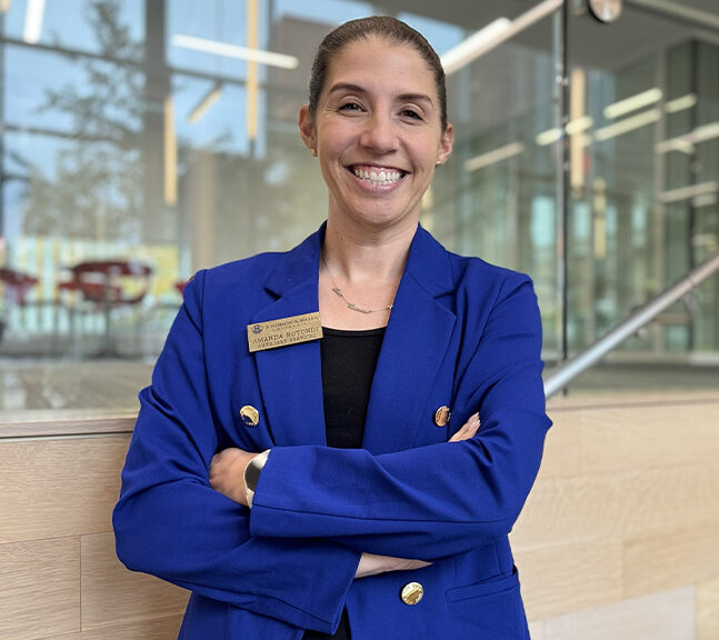 A smiling woman in a blue blazer stands with her arms crossed inside a modern building with glass walls and a staircase in the background.