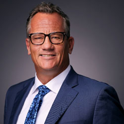 A professionally dressed individual wearing glasses and a suit with a patterned tie smiles at the camera against a neutral background.