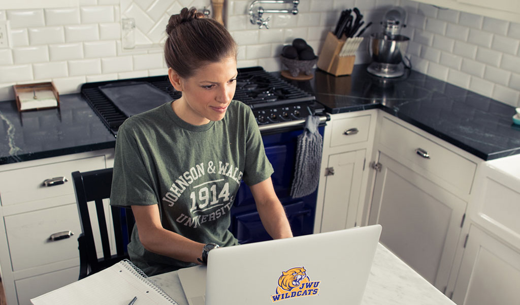 JWU Online student sitting at home in kitchen w open laptop