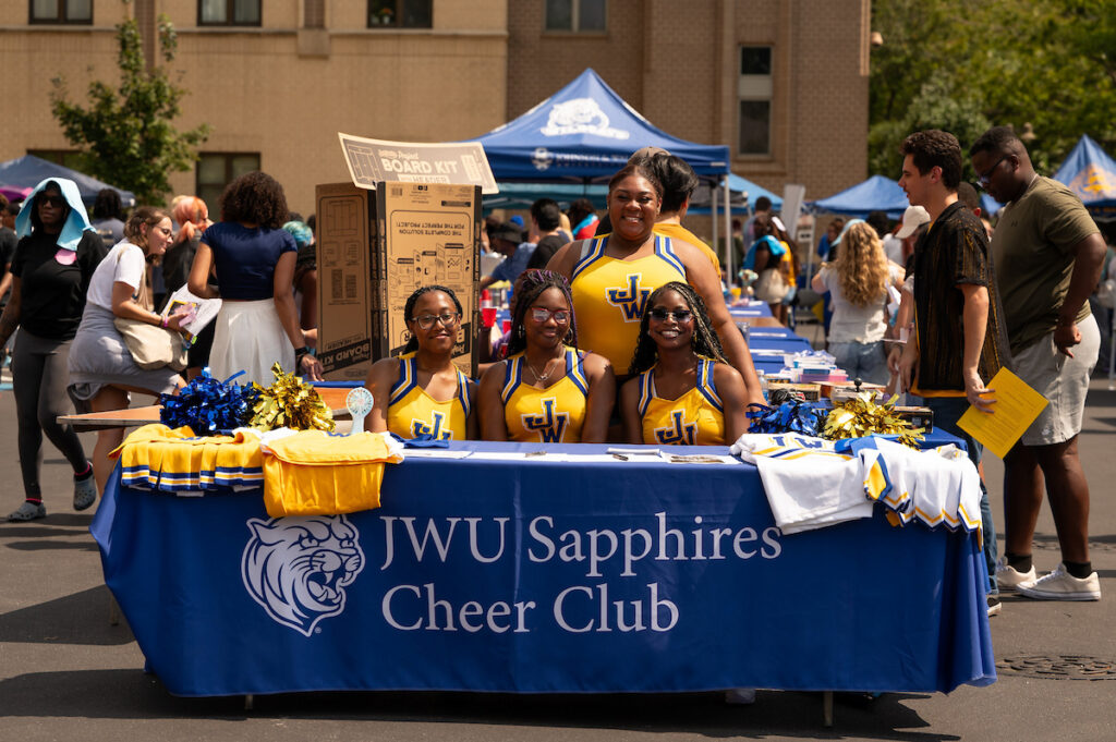 Members of the JWU Sapphires Cheer Club stand behind a table displaying team merchandise at an outdoor event.