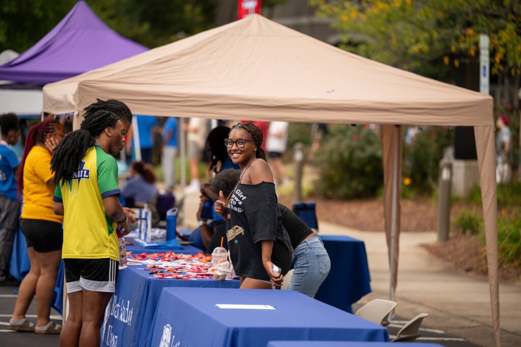 Two individuals engage at an outdoor event under a beige canopy, with one smiling and gesturing while standing beside a table covered in blue cloth and promotional items.