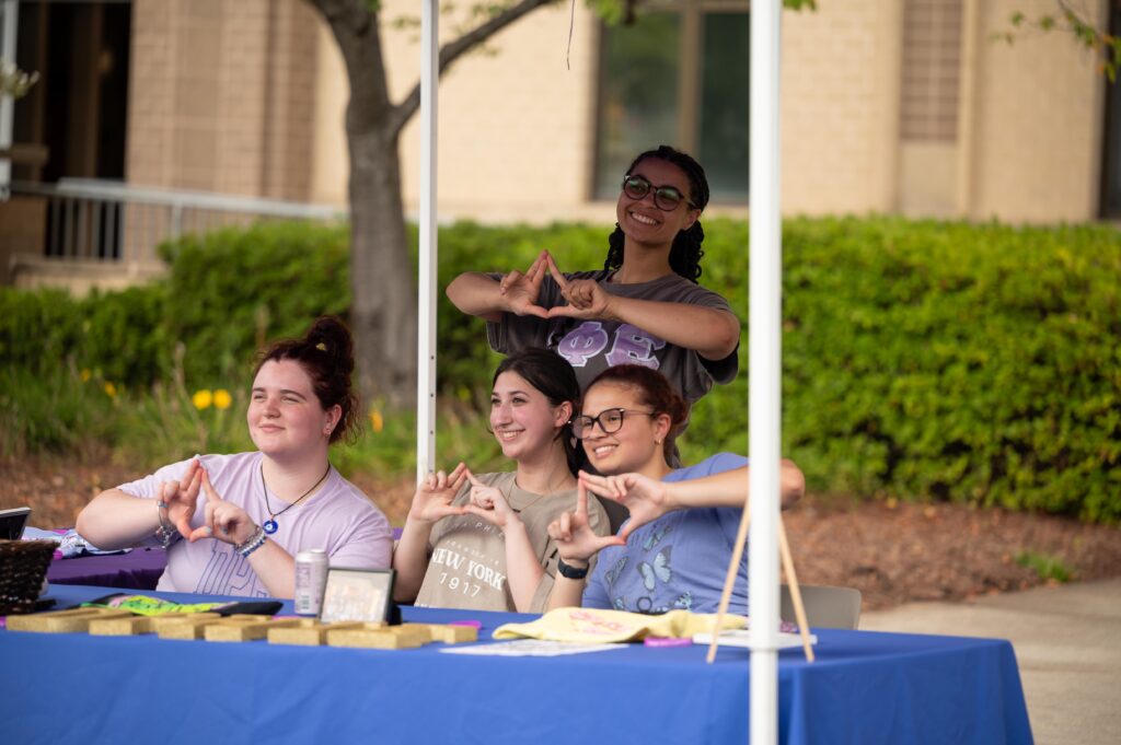 Four individuals make heart shapes with their hands while seated at a table outdoors.