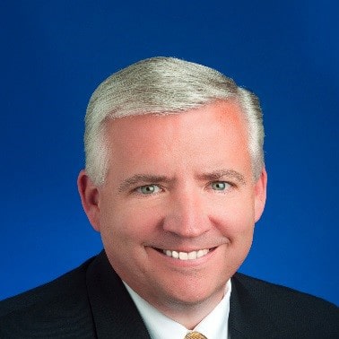 A person with gray hair and a suit smiles against a solid blue background.
