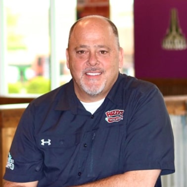A person wearing a dark blue shirt with a "Pizzeria" logo and an Under Armour emblem smiles while seated indoors.