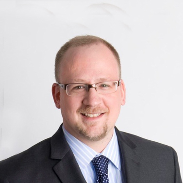 A person with glasses and a beard, wearing a suit and tie, smiles at the camera against a plain background.