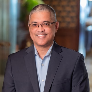 A professionally dressed individual with glasses smiles for the camera in a well-lit indoor setting.