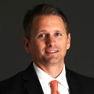 A professionally dressed individual in a dark suit and orange tie smiles against a dark background.