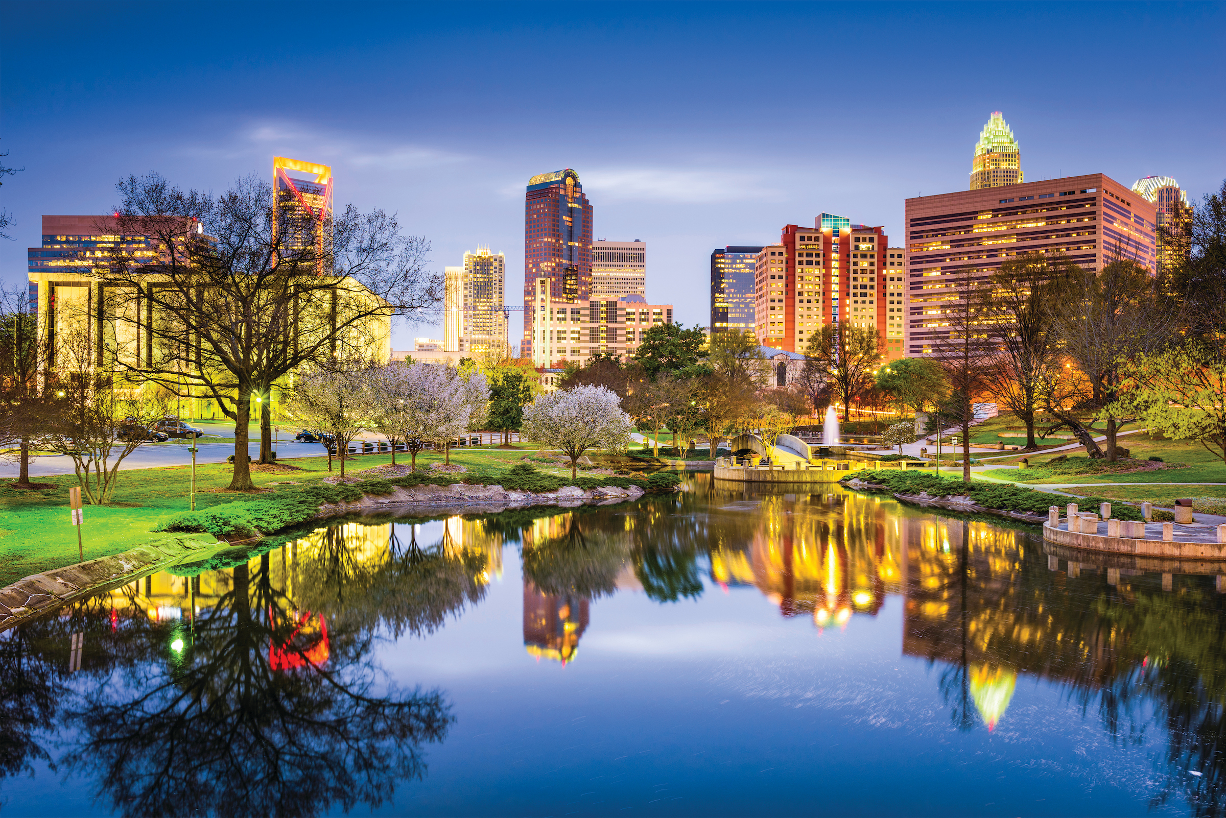 A serene urban park at dusk, with a calm pond reflecting the illuminated skyscrapers and bare trees against a deep blue sky.