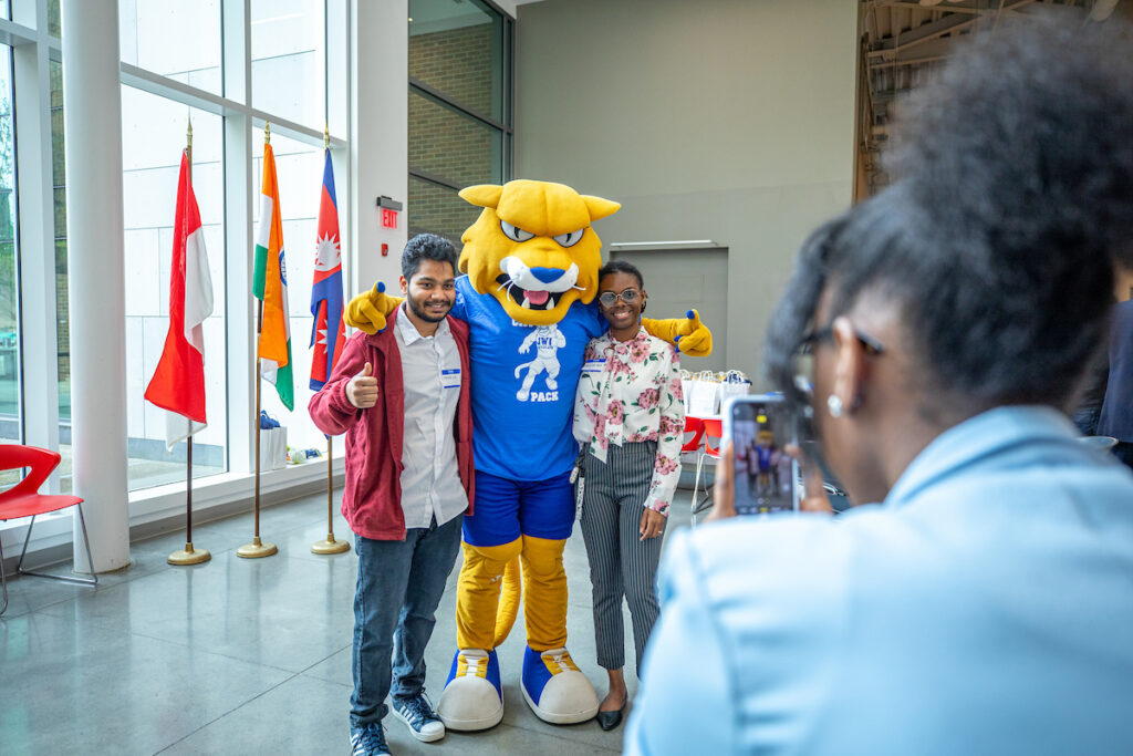 Two individuals pose with a yellow mascot in a brightly lit indoor space, while another person captures the moment on a smartphone.
