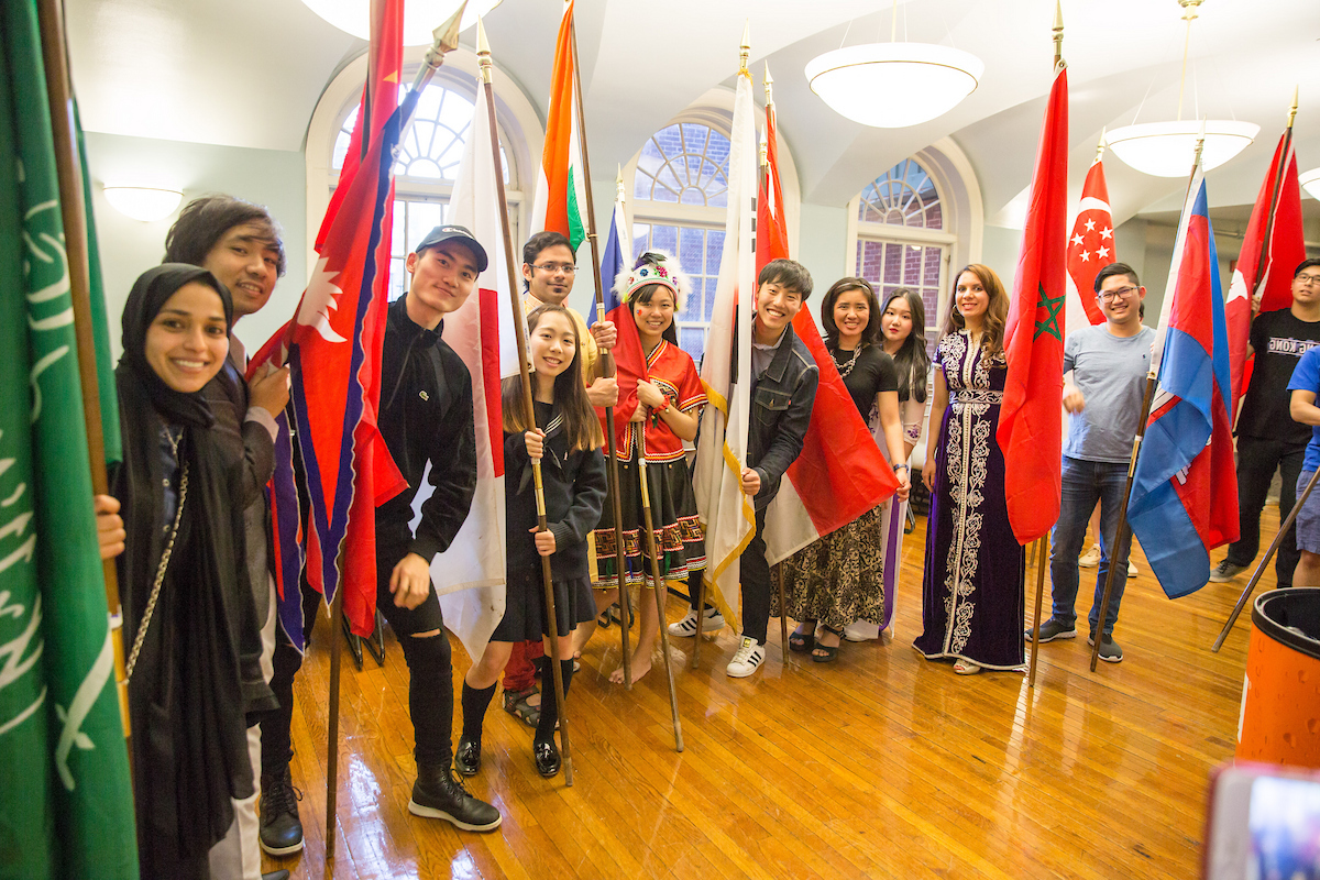 A group of diverse individuals stand together indoors, each holding a national flag, smiling for the camera in a room with arched windows and wooden floors.