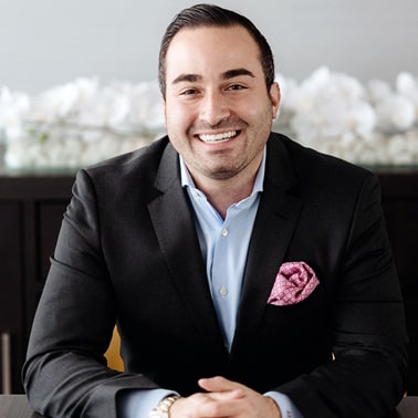 A person in a black suit with a light blue shirt and a pink pocket square smiles warmly while seated.