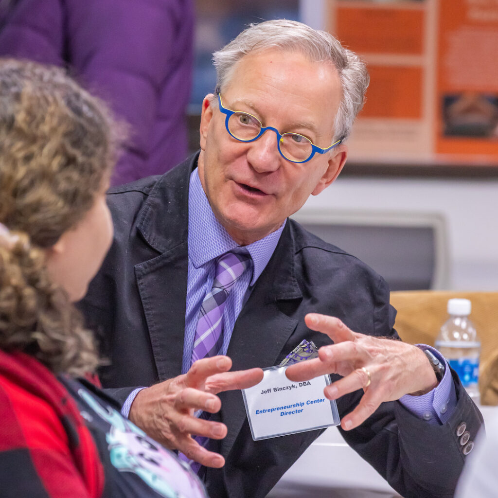 A man in a suit and blue-rimmed glasses gestures while speaking to someone at a table during an event.