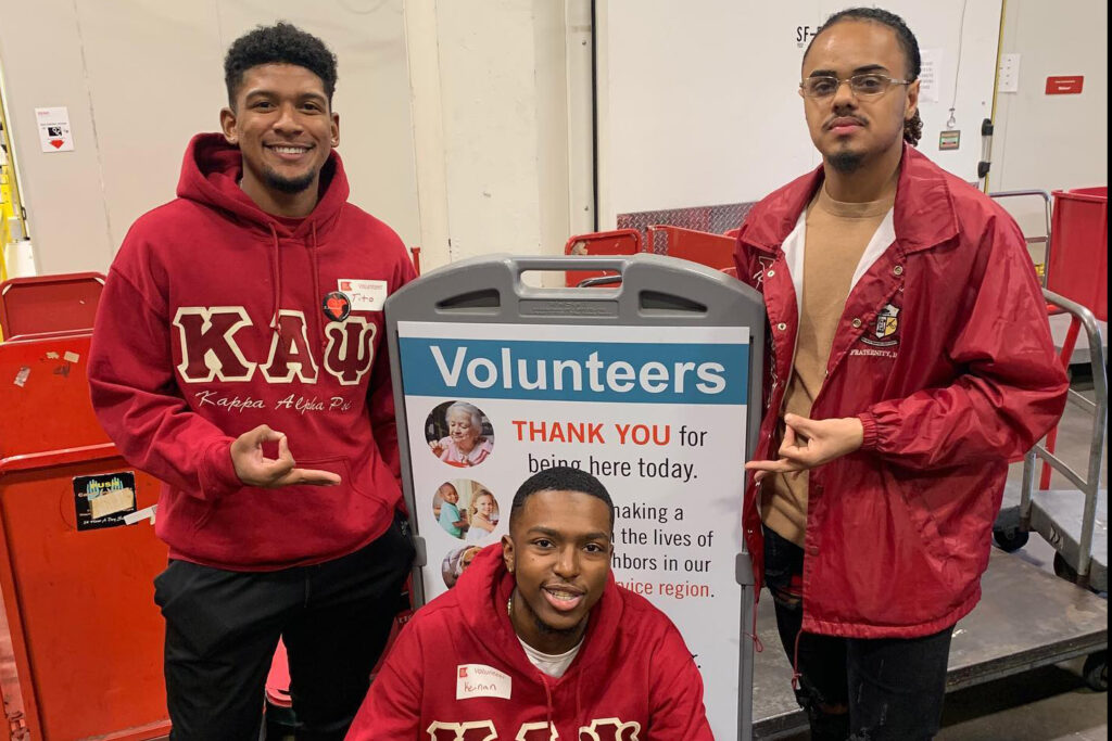 Three Johnson & Wales University students from Kappa Alpha Psi pose together while volunteering at a community event. Two students stand beside a seated student, all wearing red Kappa Alpha Psi clothing. The two standing students make hand gestures associated with the fraternity, with a volunteer sign behind them.