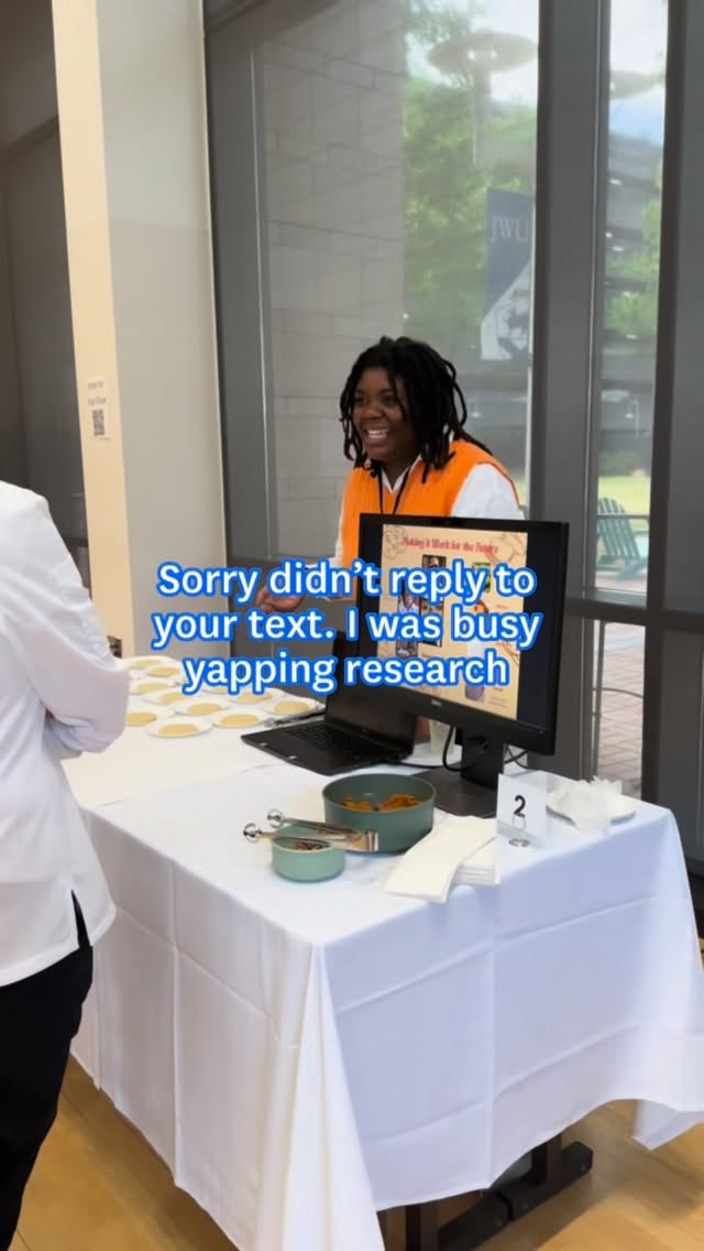 A student stands behind a research presentation table with a computer monitor and food samples. On-screen text reads, “Sorry didn’t reply to your text. I was busy yapping research.”