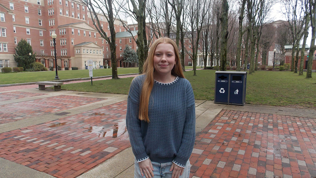 A Johnson & Wales University student stands on campus in front of brick academic buildings and a tree-lined walkway. She wears a blue sweater and jeans.