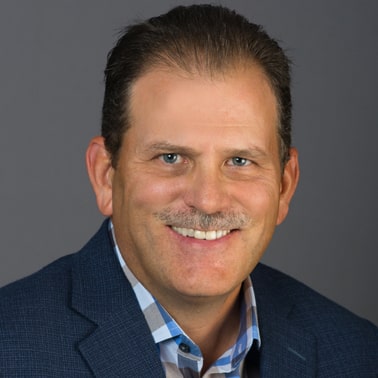 A smiling individual with a mustache, wearing a blue blazer over a patterned shirt, against a plain gray background.