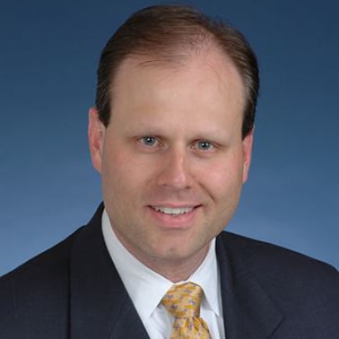 A professionally dressed individual in a suit and tie smiles against a solid blue background.